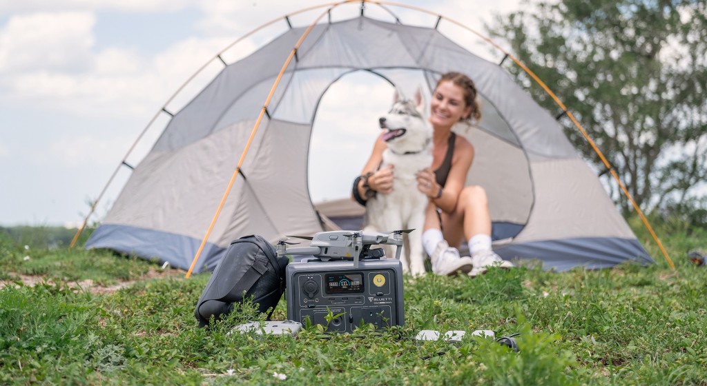 Woman and dog sitting outside a tent with a Bluetti portable power system on the grass charging a drone. Affiliated with SpookyMrsGreen.com pagan lifestyle blog.